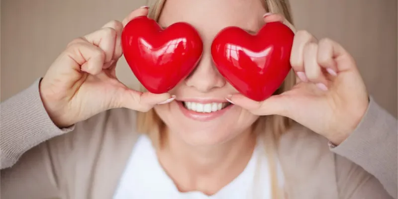 Smiling woman covering her eyes with two red heart shapes, representing joy and the cardiovascular benefits of massage.